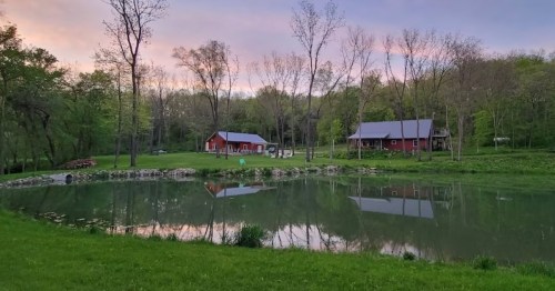 A serene landscape featuring a pond, lush greenery, and two red barns under a colorful sky at dusk.
