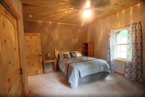 Cozy bedroom with a wooden ceiling, a bed, nightstand, and window with patterned curtains. Soft carpeted floor.