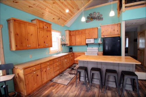 Bright kitchen with wooden cabinets, a central island with stools, and modern appliances against a turquoise wall.