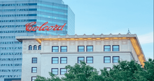 Historic building with "Colcord" sign, contrasting with modern skyscraper in the background, framed by green trees.