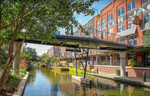 A scenic view of a canal with a bridge, lined by brick buildings and colorful boats under a clear blue sky.
