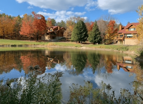 A serene pond reflects colorful autumn trees and nearby rustic buildings under a blue sky with fluffy clouds.