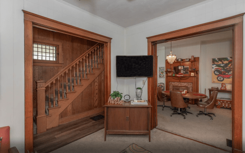 A cozy interior with wooden accents, featuring a staircase and a dining area with a round table and artwork.