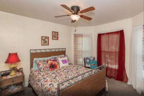 A cozy bedroom with a floral bedspread, a ceiling fan, and red curtains, featuring a nightstand with a lamp.