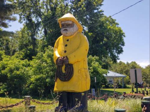 A large wooden statue of a fisherman in a yellow raincoat, holding a rope, surrounded by greenery.