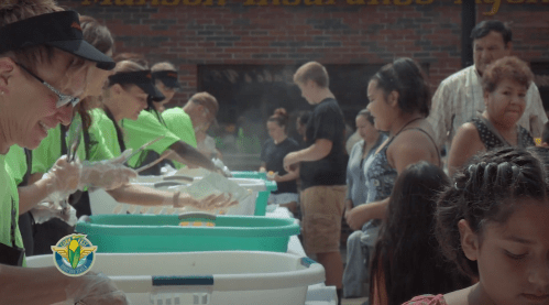 Volunteers serve food at a community event, with people lining up to receive meals in a bustling outdoor setting.