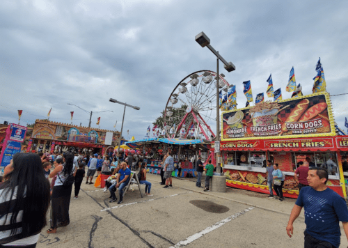 A lively fair scene with food stalls, a Ferris wheel, and crowds enjoying the festivities under a cloudy sky.