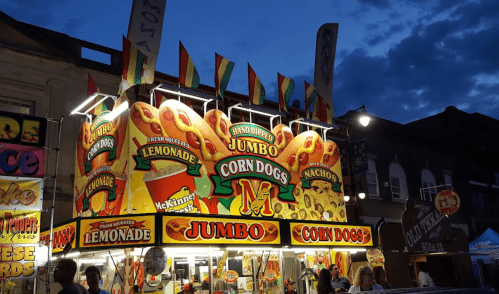 Brightly lit food stand at night, featuring signs for jumbo corn dogs, lemonade, and nachos, with colorful flags above.