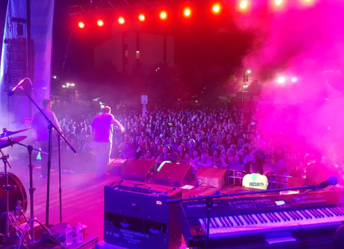 A vibrant concert scene with a large crowd enjoying a performance under colorful stage lights and smoke.