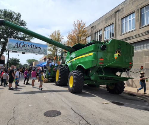 A large green combine harvester on a street, with people and a banner reading "AGtivities: A farm fun experience."