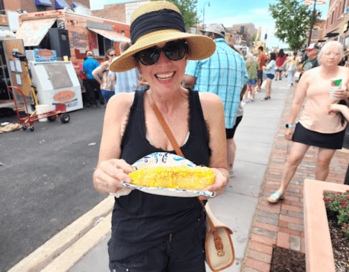 A smiling woman in a sunhat holds a plate of corn on the cob at a busy outdoor event.