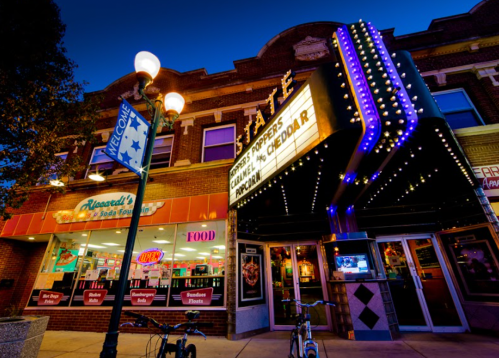 Historic theater marquee illuminated at dusk, with nearby shops and street lamps in a vibrant downtown setting.
