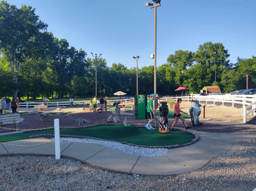 A lively mini-golf course with players, greenery, and shaded areas under a clear blue sky.