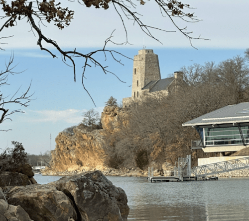 A stone tower on a rocky hill overlooks a calm lake, with a modern building nearby and trees in the foreground.