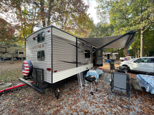 A parked travel trailer with an awning, surrounded by trees and camping gear in a serene outdoor setting.