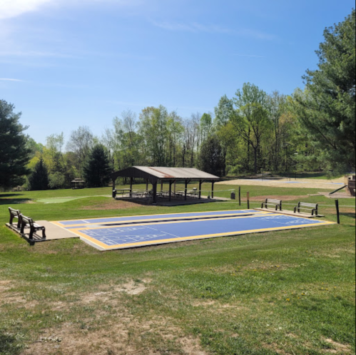 A sunny park scene featuring a large blue court, benches, and a covered pavilion surrounded by trees.