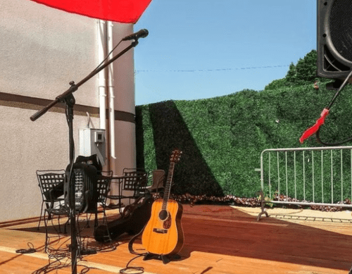 A stage setup with a microphone, guitar, and chairs, surrounded by greenery and a clear blue sky.