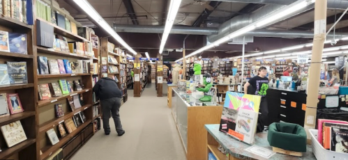 A spacious bookstore with wooden shelves filled with books, customers browsing, and a checkout area in the foreground.
