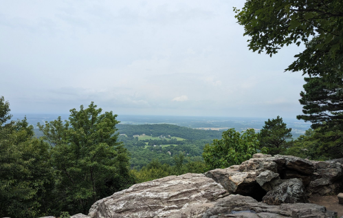A scenic view from a rocky overlook, featuring lush green trees and rolling hills under a cloudy sky.