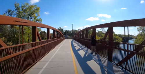 A person stands on a pedestrian bridge with metal arches, overlooking a river under a clear blue sky.
