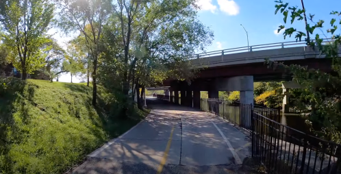 A paved path runs under a bridge, surrounded by trees and grass, with a river visible on the right.
