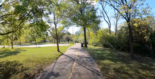 A paved path through a park lined with trees and greenery, with sunlight filtering through the leaves.