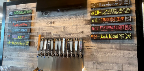 A wooden wall displaying a variety of craft beers on tap, with a chalkboard menu listing beer names and ABV percentages.