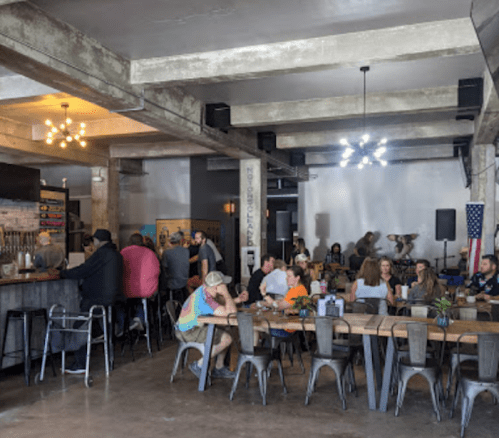 A lively bar and dining area with patrons enjoying food and drinks, featuring industrial decor and a stage in the background.