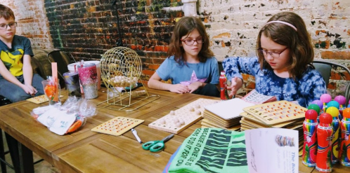 Two children sit at a table with bingo cards and craft supplies, focused on a game in a rustic setting.
