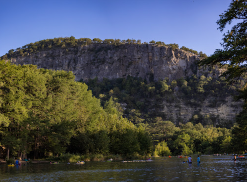 A serene river scene with a steep cliff and lush greenery, featuring people enjoying the water.