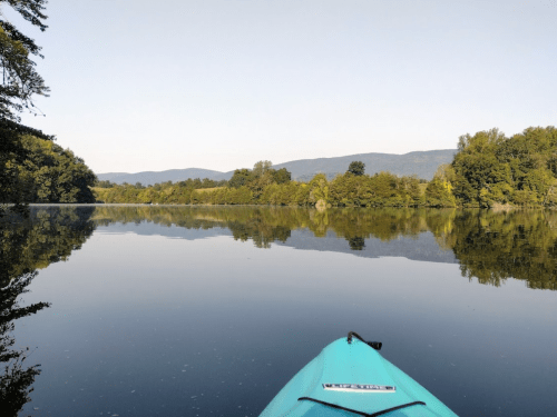 A serene view from a kayak on calm water, reflecting trees and mountains under a clear sky.