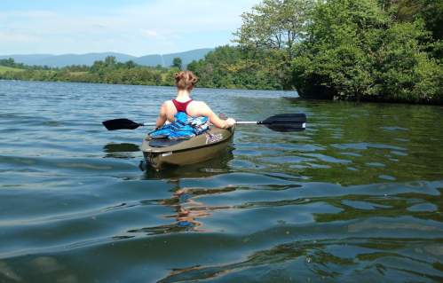 A person in a kayak paddles on a calm lake surrounded by trees and mountains on a sunny day.