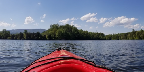 A red kayak on calm water, with trees and mountains in the background under a clear blue sky with fluffy clouds.