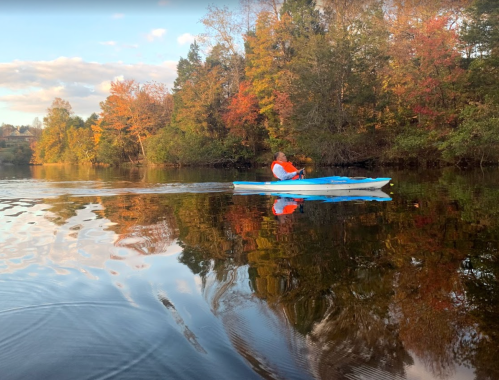 A person in a kayak paddles on a calm lake, surrounded by trees with vibrant autumn foliage reflecting in the water.