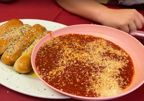 A pink bowl of marinara sauce with grated cheese, accompanied by three garlic breadsticks on a plate.