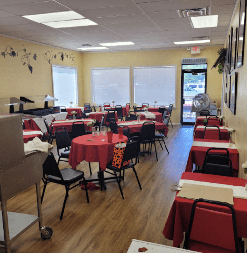 A cozy restaurant interior with red tablecloths, black chairs, and a fan, featuring a warm, inviting atmosphere.