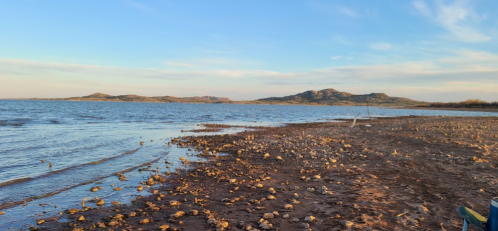 A serene lakeside view with rocky shorelines and distant hills under a clear blue sky.