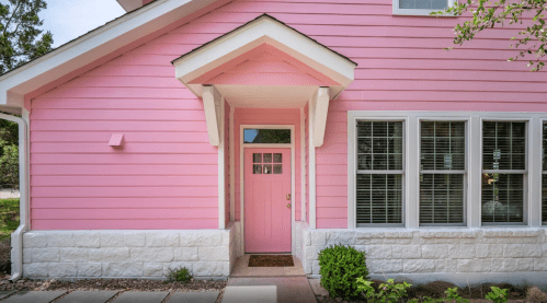 A charming pink house with a matching front door and large windows, surrounded by greenery and a stone pathway.