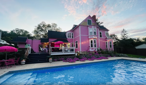 A vibrant pink house with a pool, surrounded by flowers and outdoor seating, under a colorful sky at sunset.