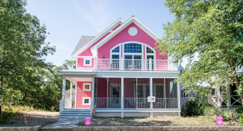 A large pink house with white trim, featuring a spacious porch and large windows, surrounded by trees.
