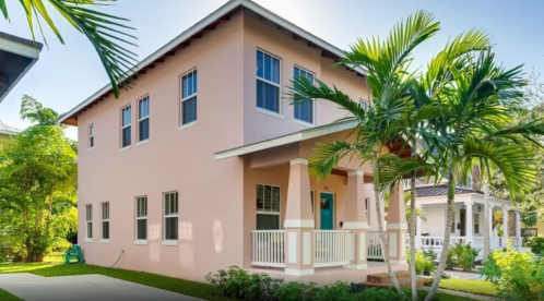 A two-story pink house surrounded by palm trees and greenery, featuring a front porch and blue door.