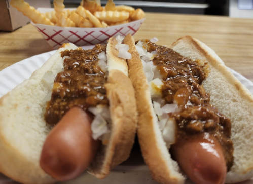 Two hot dogs in buns topped with chili and onions, served with a side of crinkle-cut fries in a basket.
