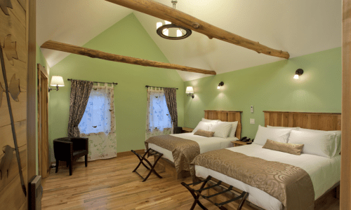 Cozy hotel room with two beds, wooden accents, green walls, and natural light from windows.
