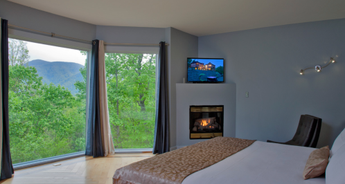 A modern bedroom with a large window overlooking greenery, a TV on the wall, and a cozy fireplace.