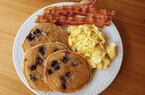 A plate with three blueberry pancakes, scrambled eggs, and crispy bacon strips on a wooden table.