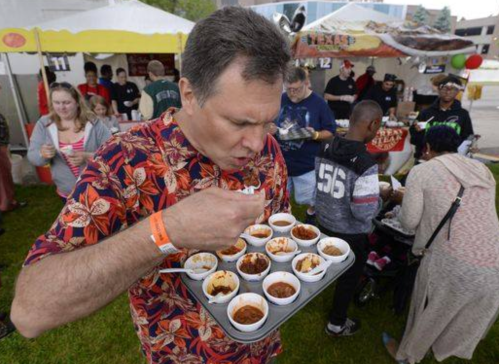 A man in a floral shirt samples food from small cups at an outdoor festival, surrounded by people and tents.