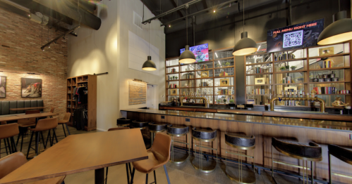 A modern bar interior with wooden shelves, high stools, and a well-stocked counter, featuring warm lighting and brick walls.