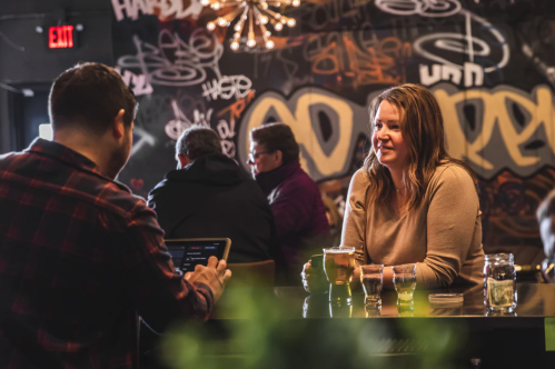 A woman sits at a bar, smiling at a man using a tablet, with a graffiti-covered wall in the background.