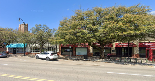 Street view featuring shops with colorful awnings, trees lining the sidewalk, and a clear blue sky.
