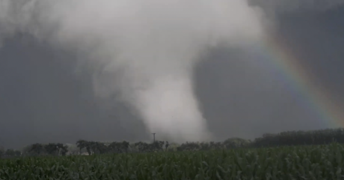 A tornado touches down in a field, with a rainbow visible in the background against a stormy sky.